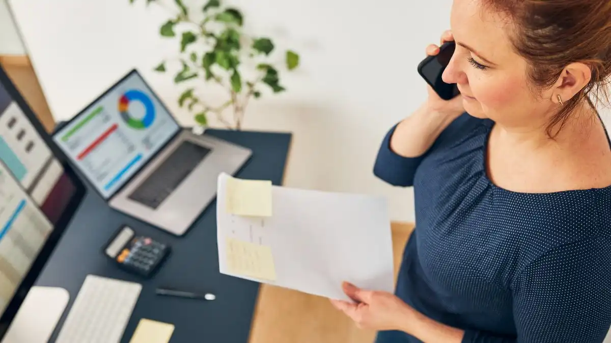 Mulher em ambiente de trabalho utilizando telefone e analisando gráficos em computador para analisar o desconto de duplicatas