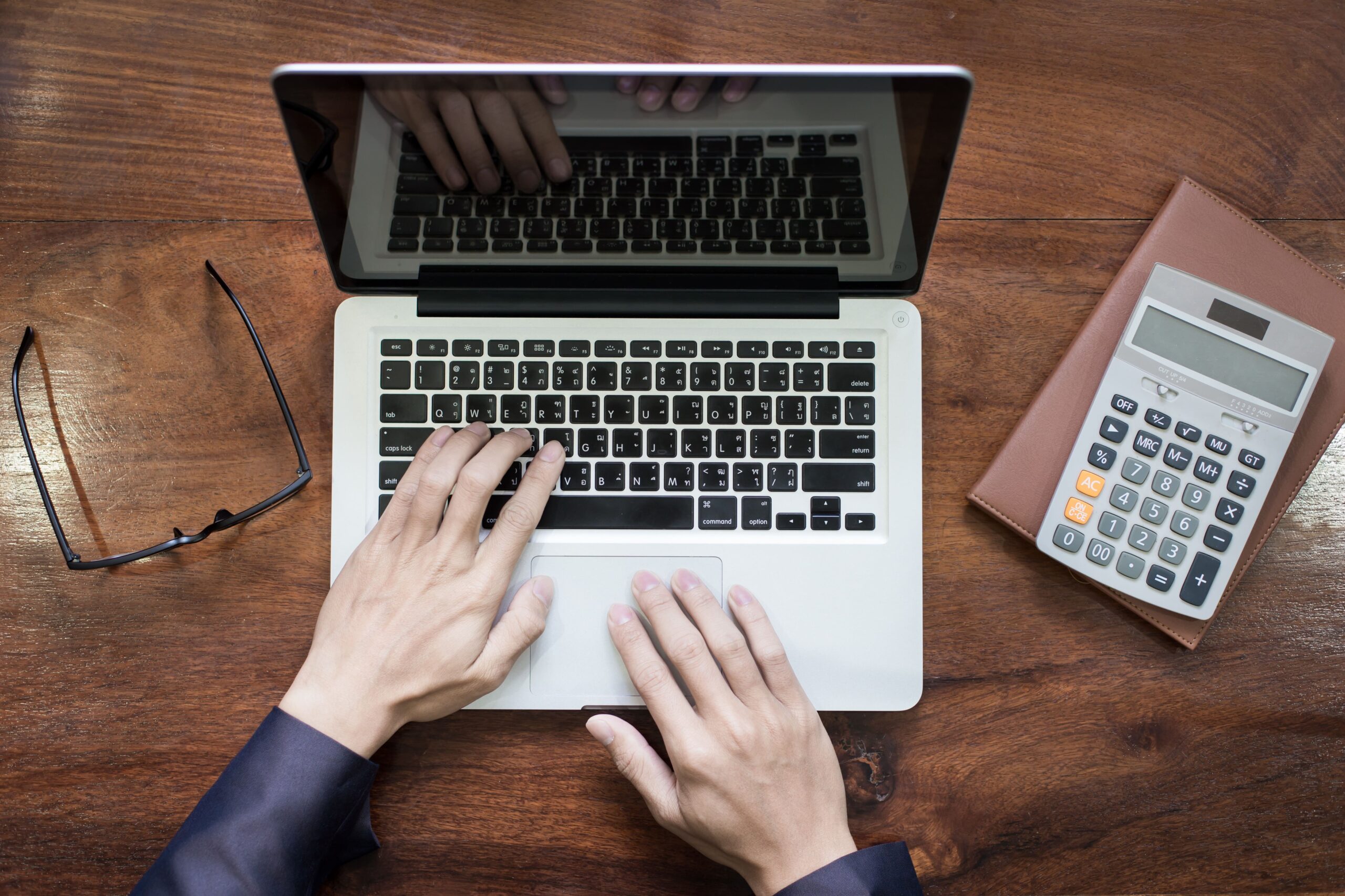 Pessoa usando laptop, calculadora e óculos de leitura sobre mesa de madeira, representando trabalho ou estudo econômico e financeiro.